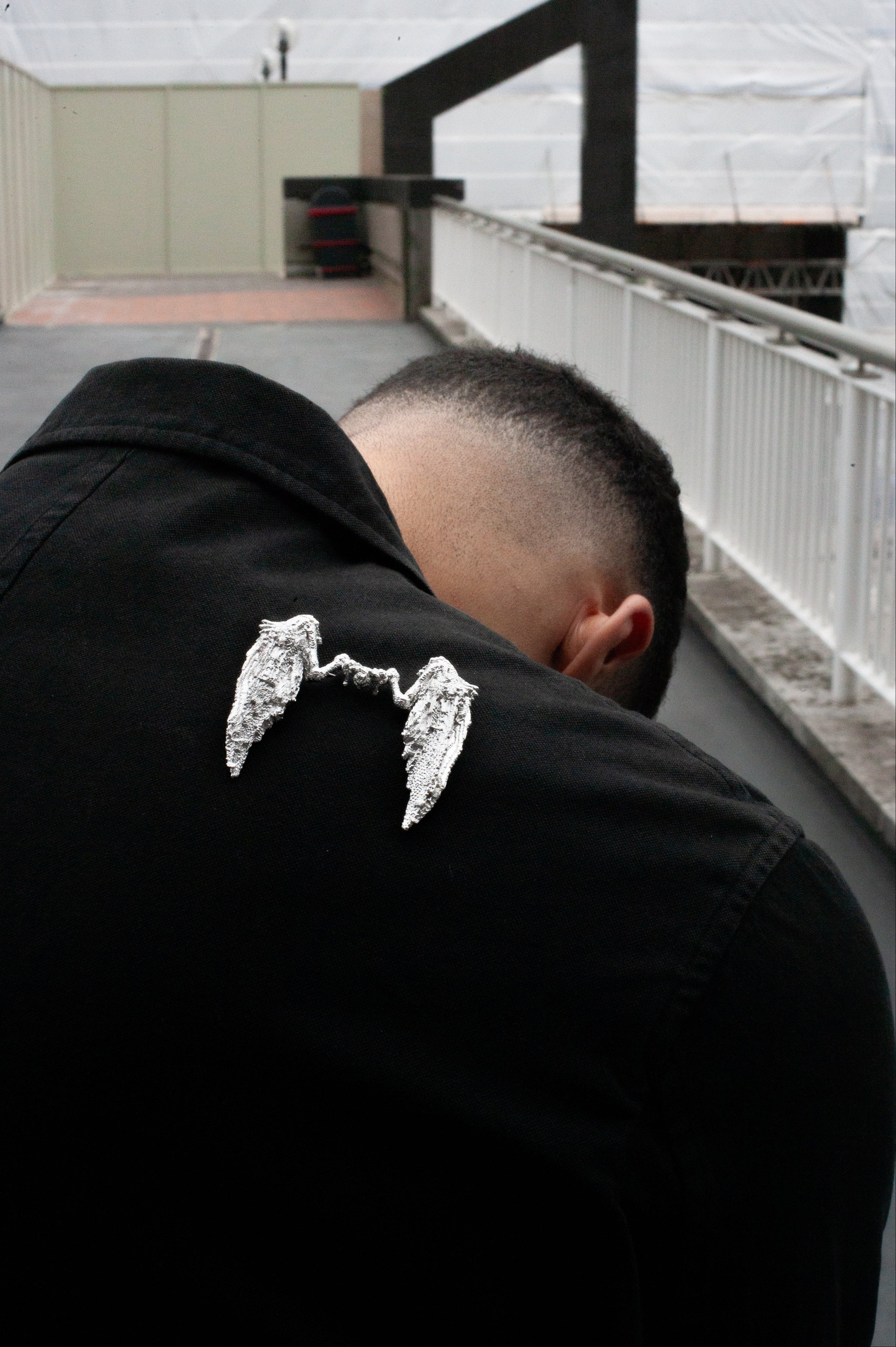 Man wearing a black jacket with a silver wings brooch on the back, standing on a bridge.